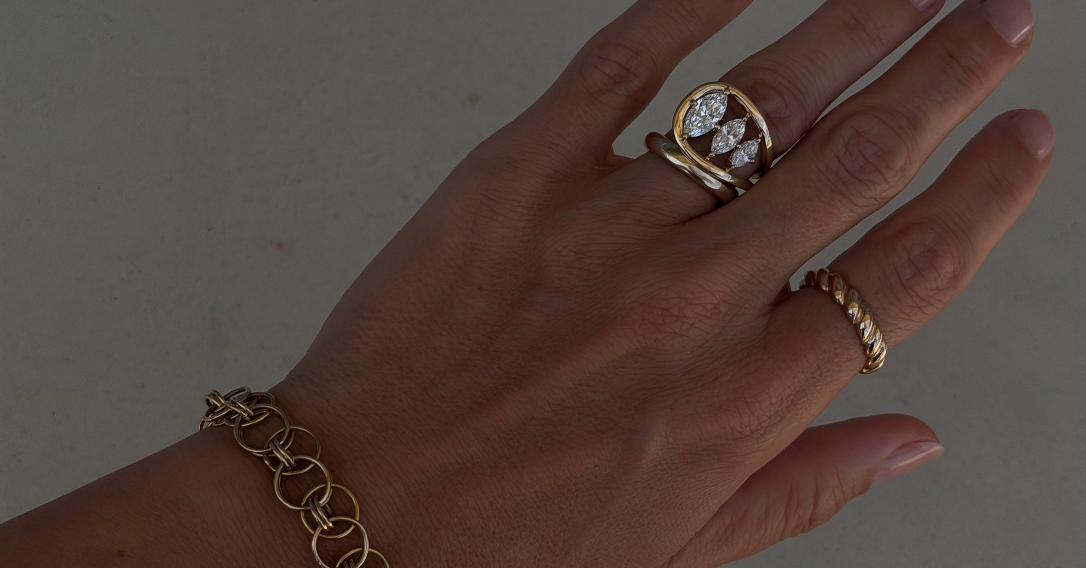 Hand wearing custom 14k gold jewelry including a custom mixed link bracelet, engagement ring with three marquise of descending sizes, a half round gold band, and a rope textured ring.
