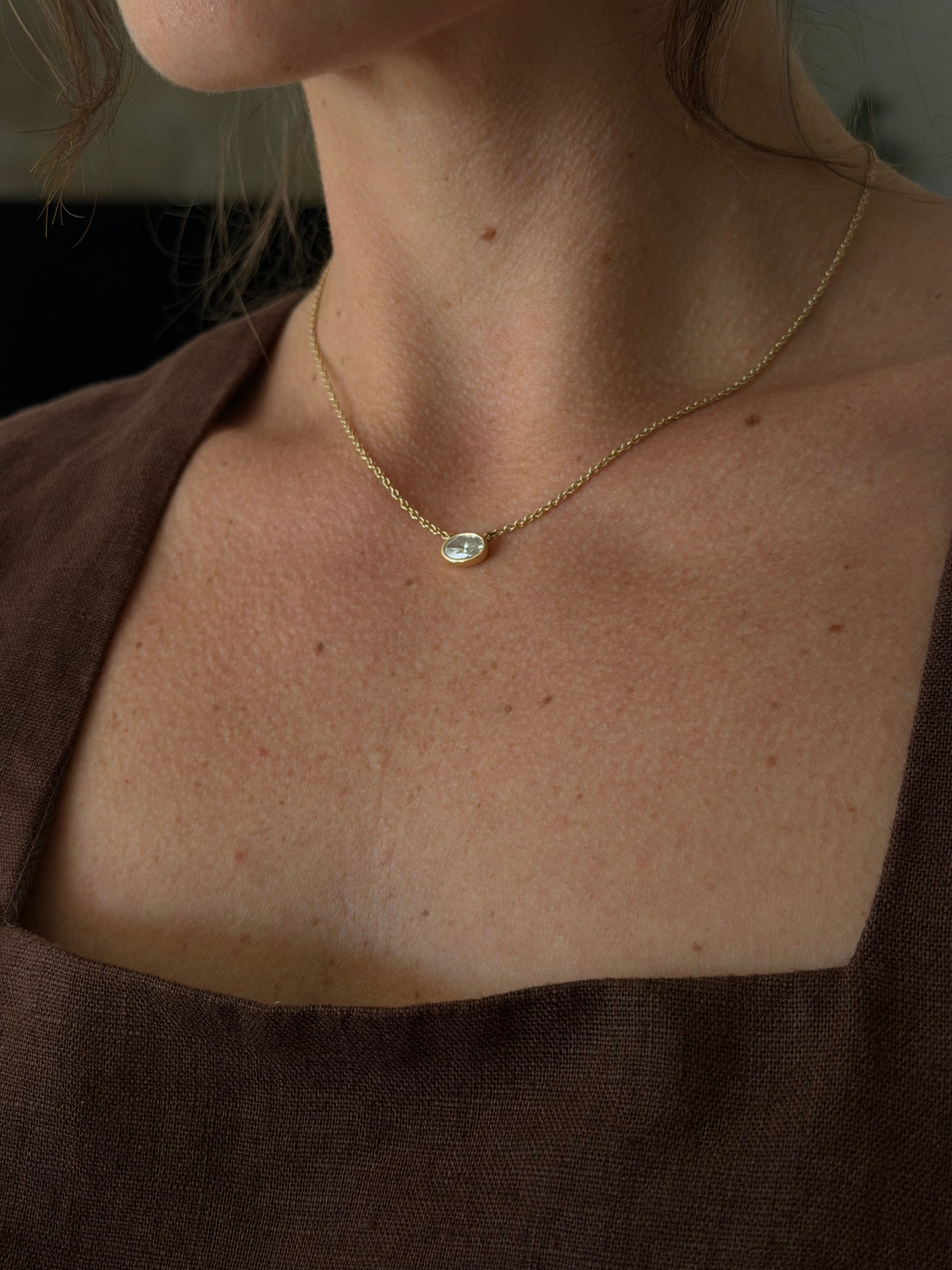 Necklace with a pendant worn by a person, focusing on the necklace which is an oval cut, 1 carat diamond, set in a 14k yellow gold bezel that is permanently attached to a gold chain.