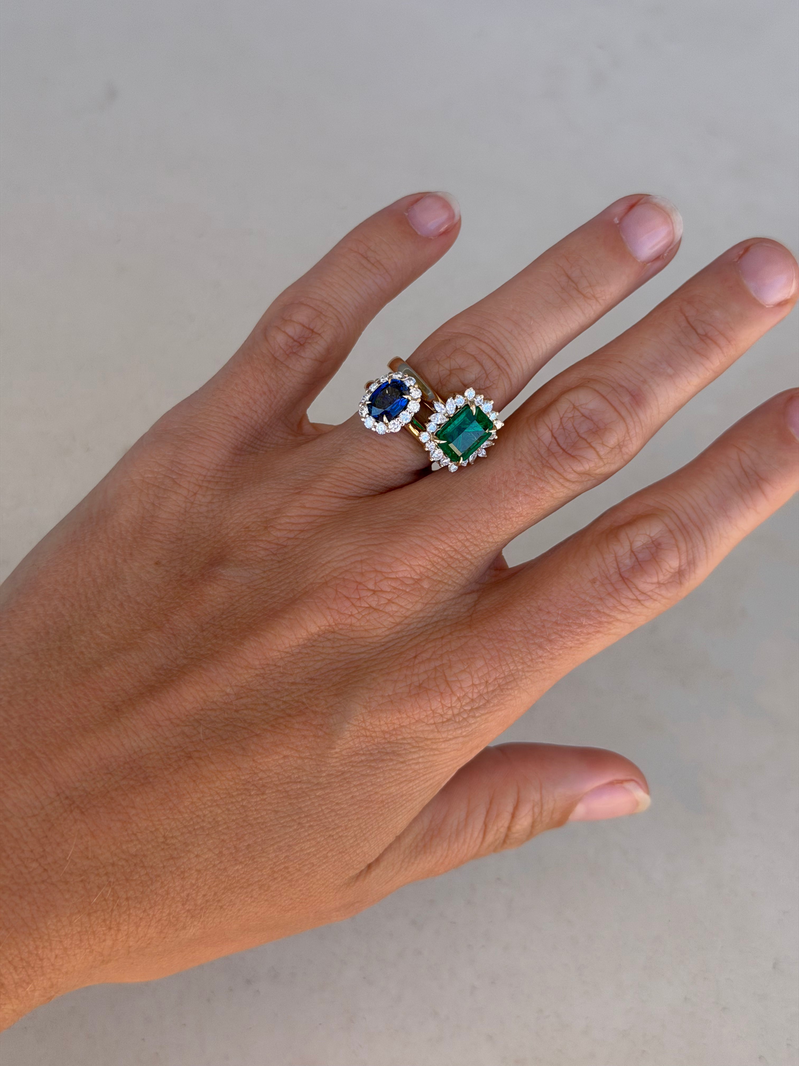 Hand wearing two rings with gemstones on a neutral background. One ring is an oval sapphire framed with diamonds like princess Diana's ring. The other is an emerald cut emerald with a frame of diamonds in various shapes like rounds and marquises.