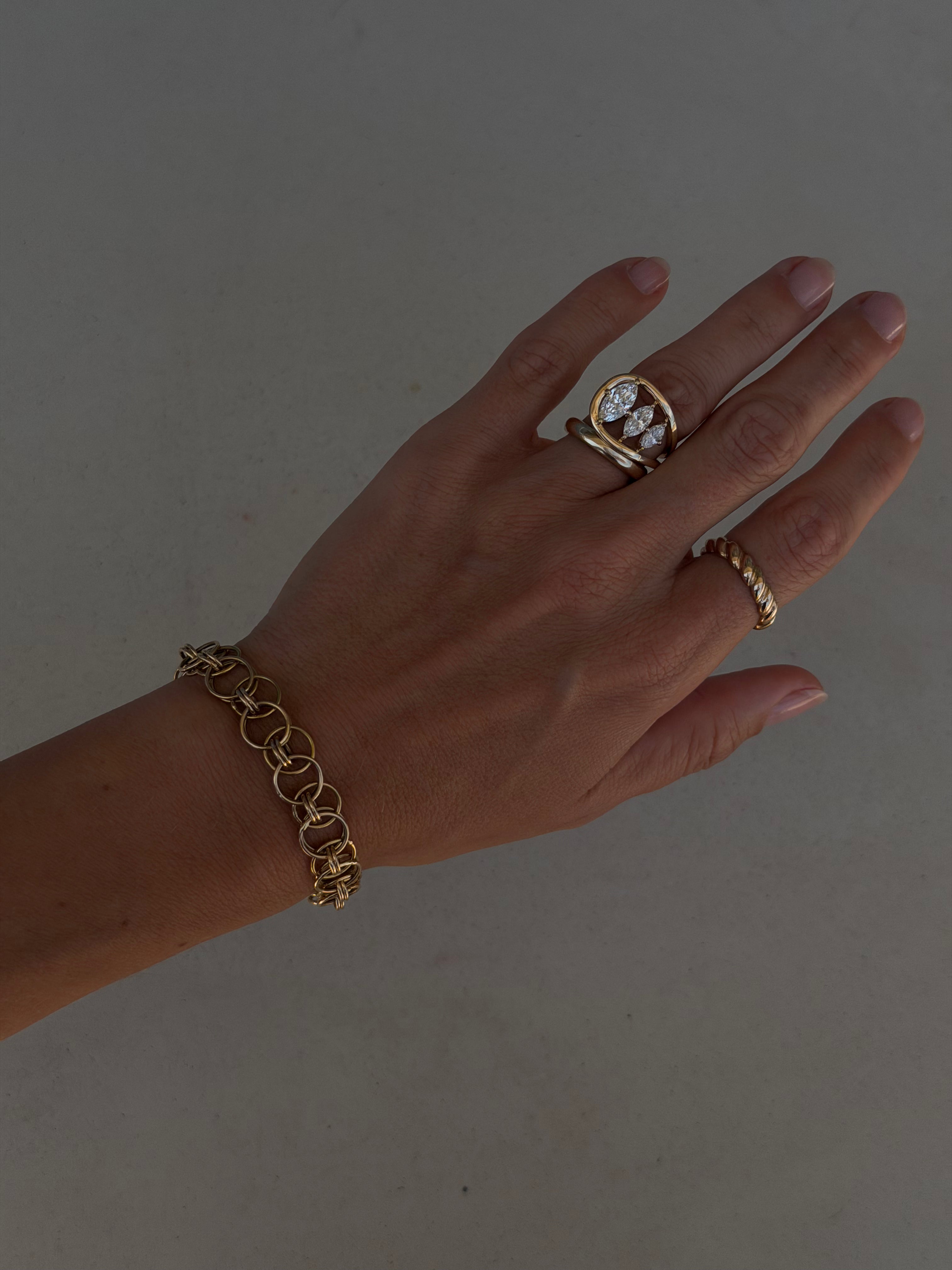 Hand wearing custom gold chain bracelet, engagement ring with asymmetrical design and three marquises descending in size, a gold band paired with the engagement ring, and a rope textured ring on the pointer finger. 