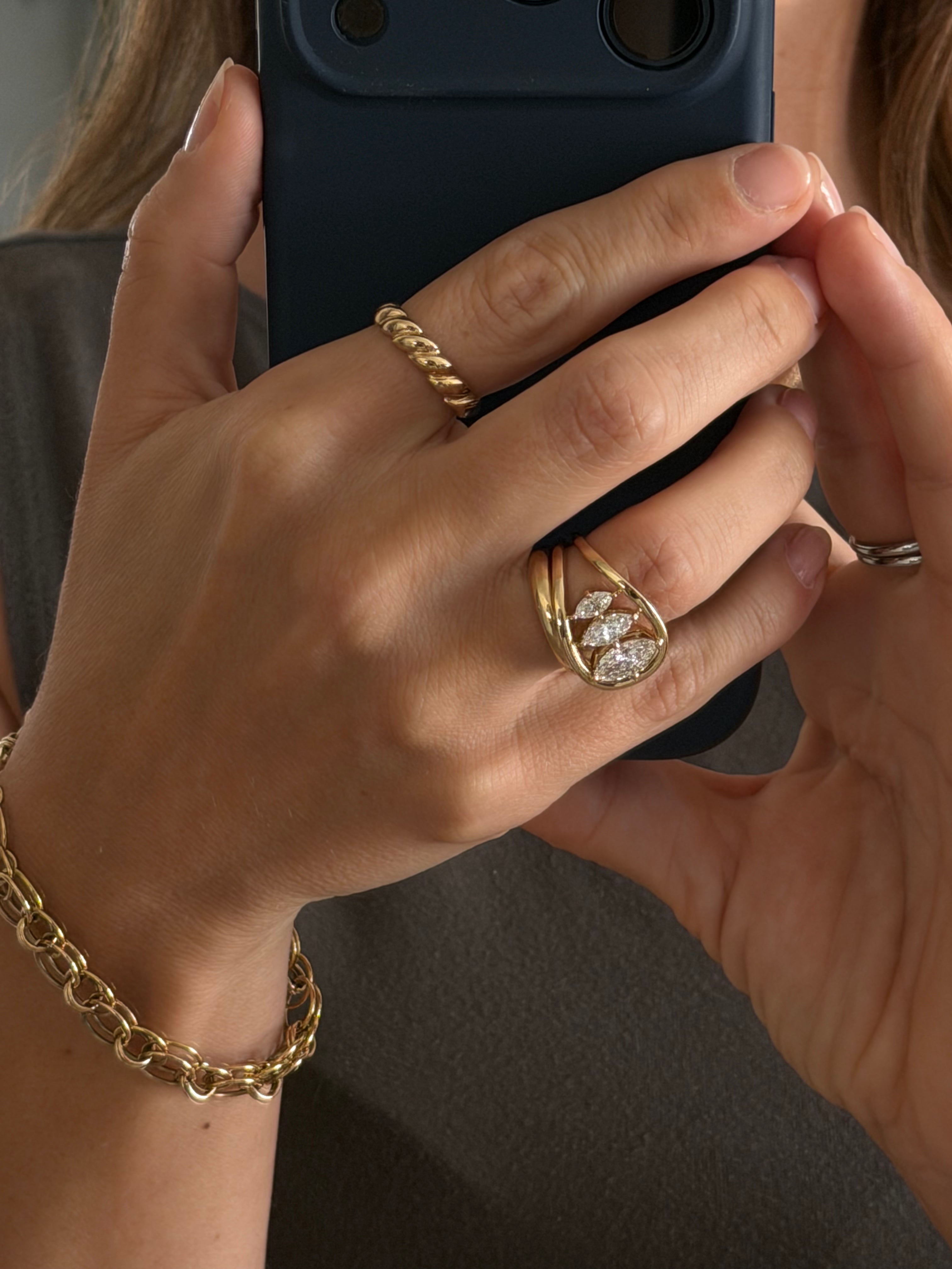 Close-up of hands holding a phone with gold jewelry on fingers and wrists including a custom gold chain, a unique asymmetrical engagement ring, a rope textured ring, and a simple half round gold band paired with the engagement ring.