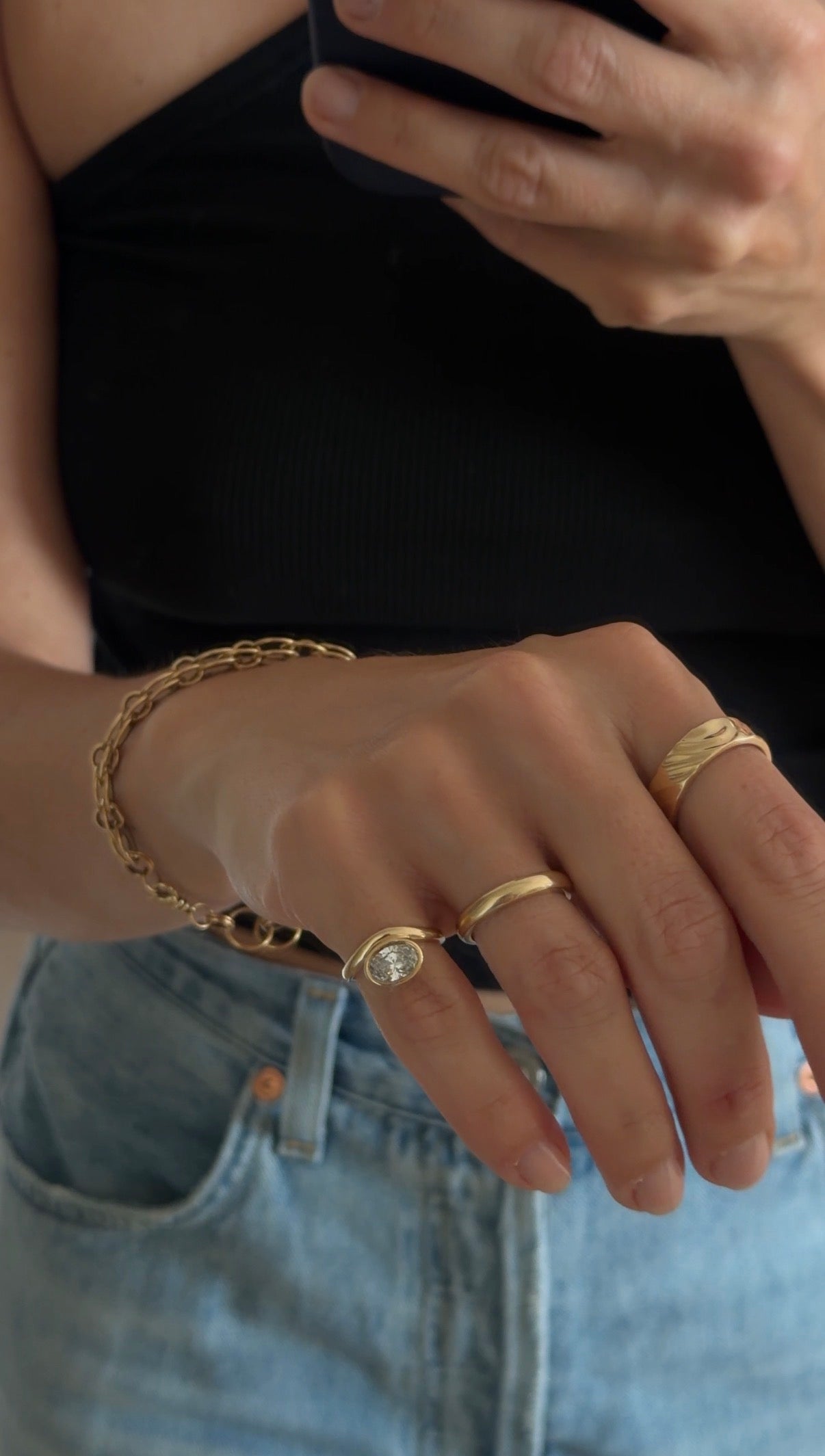 mirror selfie focused on the hand wearing a custom gold link chain, custom wave textured gold ring, a half round gold band, and a pinky ring with an organic curved ring around a bezel 1ct diamond. 
