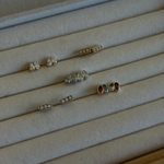Six pairs of stud earrings displayed on a beige velvet jewelry organizer tray. The earrings include silver and gold metal settings with clear stones, white stones, and colorful red, blue, and green accents.