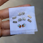 A hand holding a small stack of white tissue paper with multiple pairs of stud earrings arranged on it. The earrings include silver and gold metal settings with diamonds and gemstones.